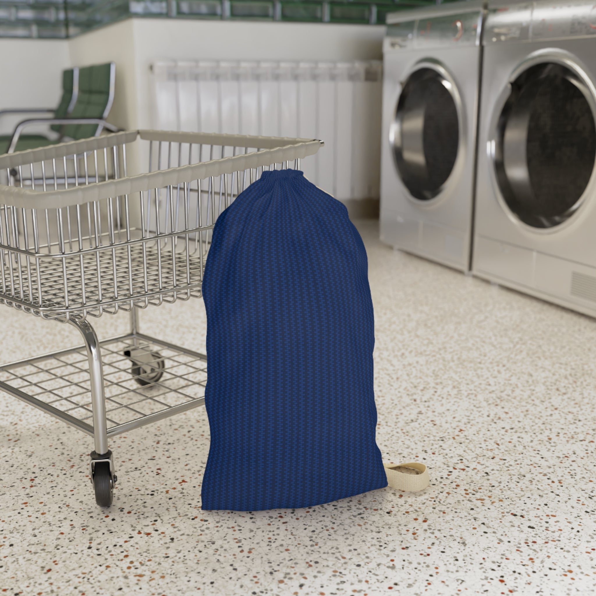 A navy blue textured drawstring laundry bag standing on a speckled floor next to a metal shopping cart in a laundry room with washing machines in the background.