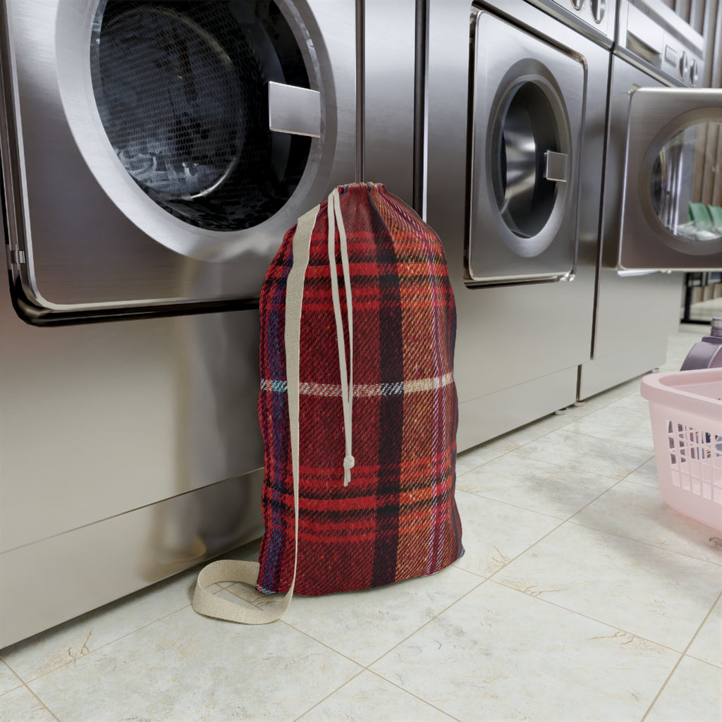 A red and black plaid drawstring laundry bag standing on a tiled floor in front of stainless steel washing machines.