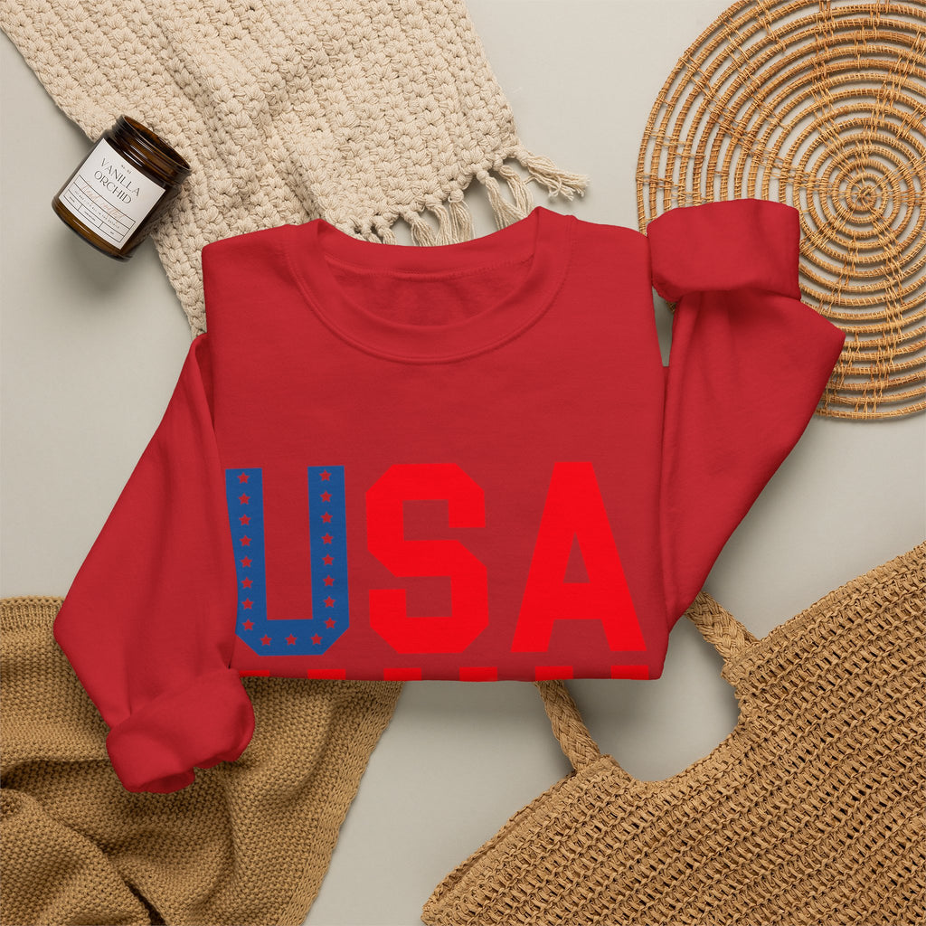 A folded red crewneck sweatshirt with a large 'USA' graphic across the chest in red and blue, styled in a flat lay with a woven blanket, a small candle jar, and a wicker tray on a light gray background.