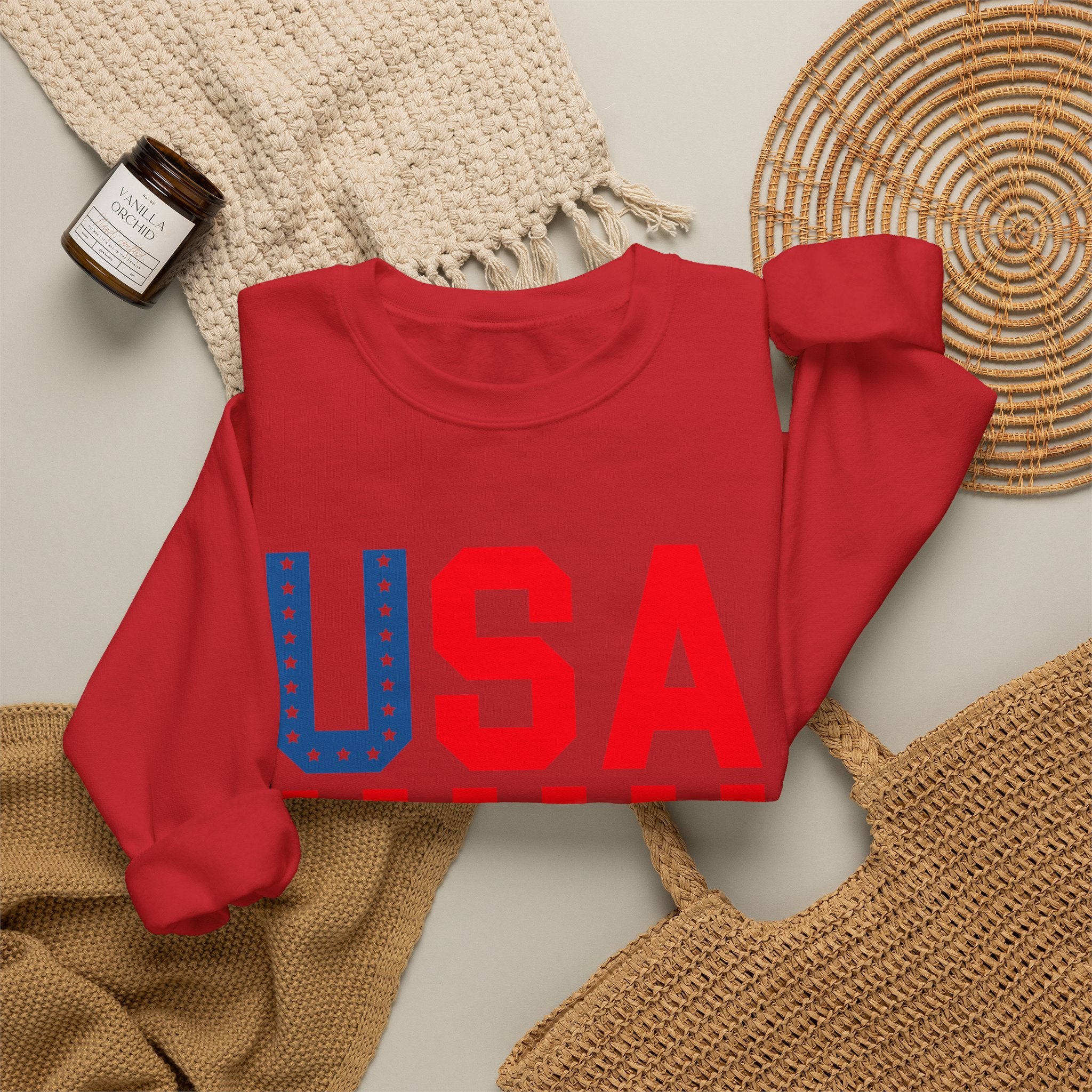 A folded red crewneck sweatshirt with a large 'USA' graphic across the chest in red and blue, styled in a flat lay with a woven blanket, a small candle jar, and a wicker tray on a light gray background.