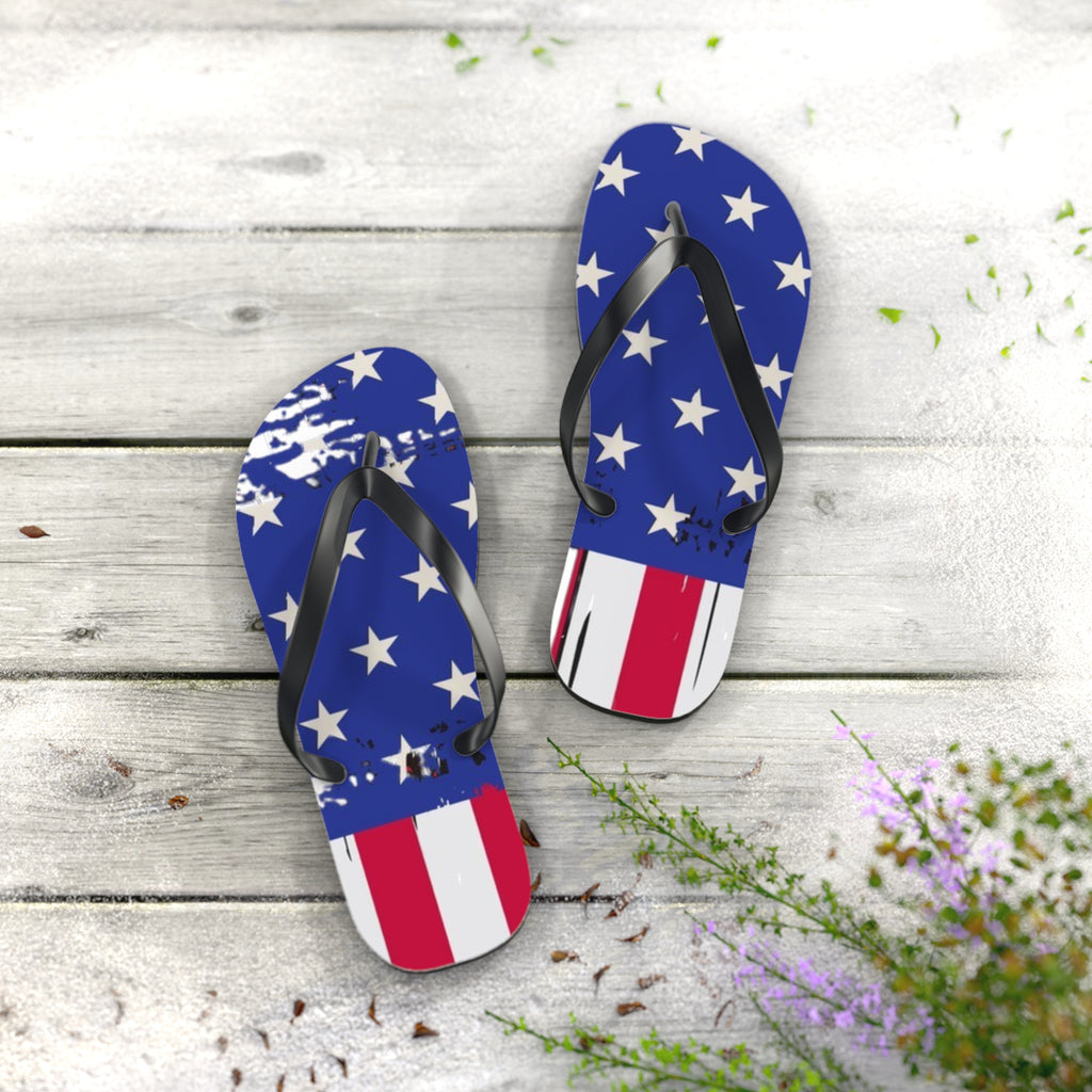 A pair of flip-flop sandals with a distressed American flag design, featuring blue straps and footbeds with white stars, and red and white stripes at the toe. The sandals are displayed on a light gray wooden plank surface with decorative green and purple wildflowers in the foreground.