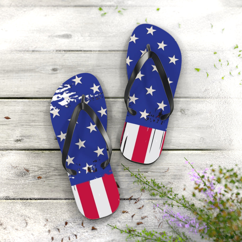 A pair of flip-flop sandals with a distressed American flag design, featuring blue straps with white stars on one side and red and white stripes on the other, displayed on a light gray wooden plank surface with decorative greenery.