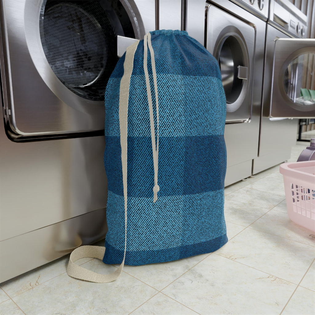 A blue and teal plaid laundry bag with a beige drawstring, standing on a tiled floor in front of stainless steel washing machines.