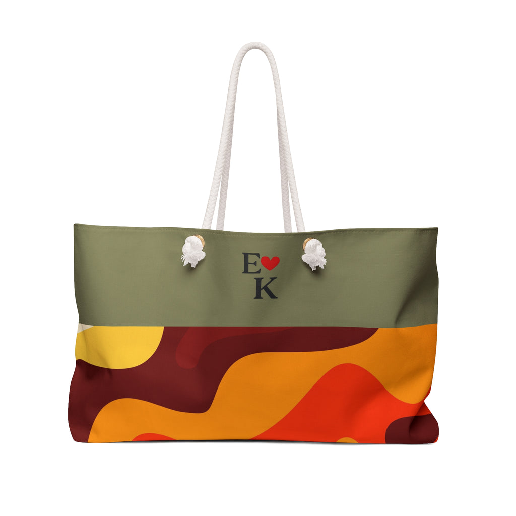 A tote bag with a two-tone design: an olive green top section featuring the text 'E [red heart] K' and two white pom-pom details, and a lower section with an abstract wavy pattern in orange, yellow, and dark red. It has white rope handles and is shown on a plain white background.