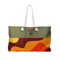 A tote bag with a two-tone design: an olive green top section featuring the text 'E [red heart] K' and two white pom-pom details, and a lower section with an abstract wavy pattern in orange, yellow, and dark red. It has white rope handles and is shown on a plain white background.