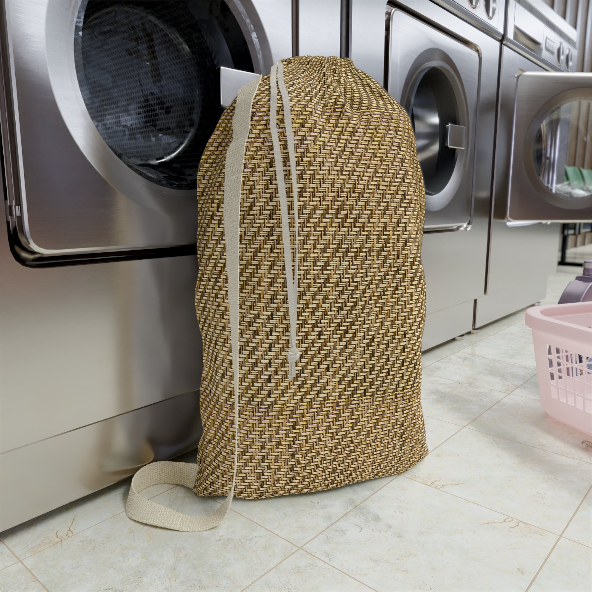 A woven beige and brown drawstring laundry bag standing upright in front of stainless steel washing machines on a tiled floor.