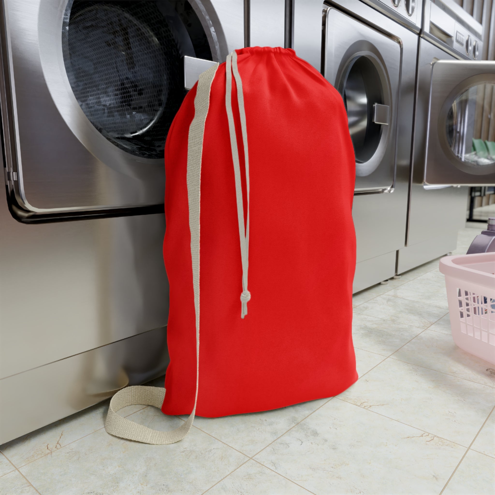 A bright red drawstring laundry bag with a beige shoulder strap and drawstring, standing upright on a tiled floor in front of stainless steel washing machines.