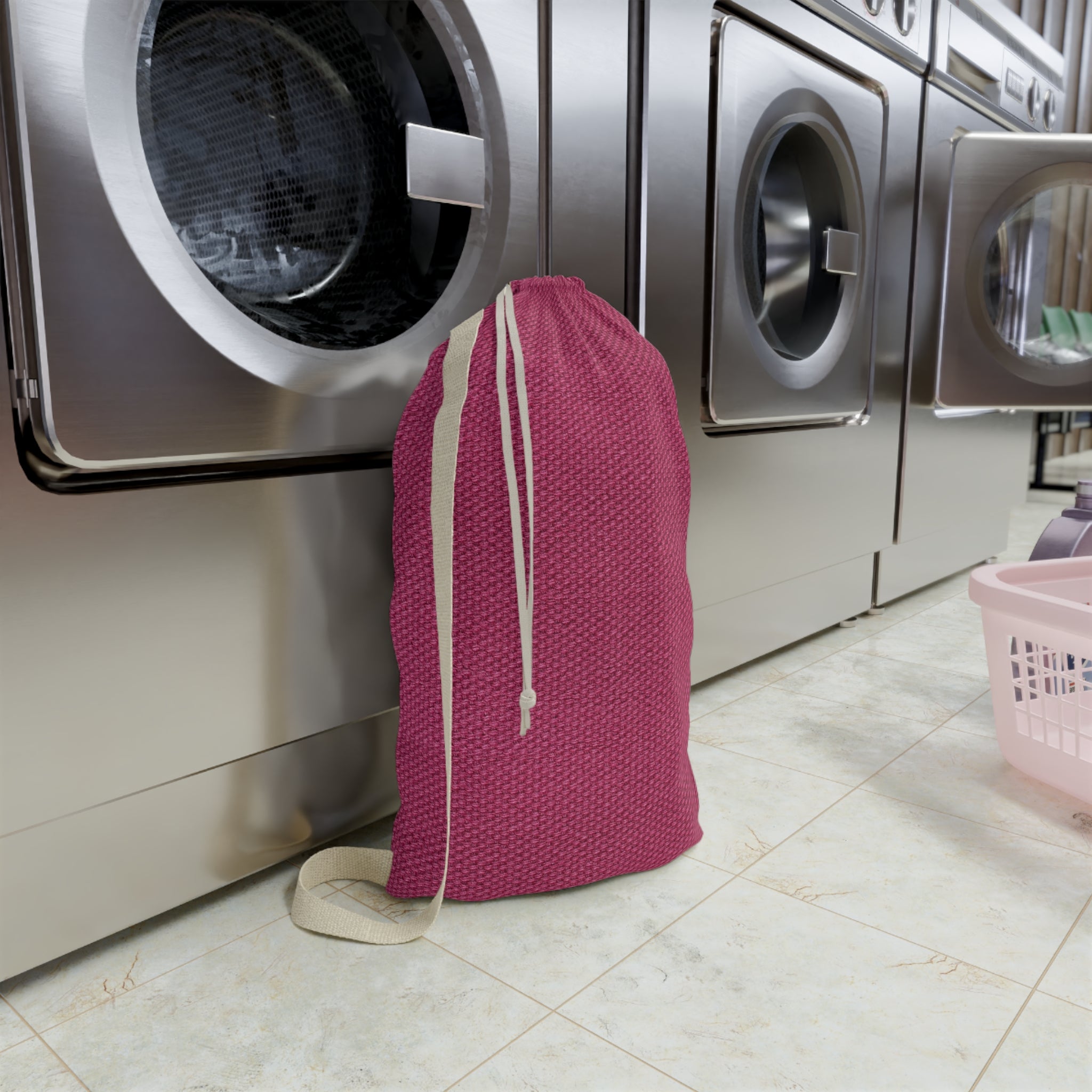 A pink drawstring laundry bag with a textured pattern stands on a tiled floor in front of stainless steel washing machines in a laundry room.