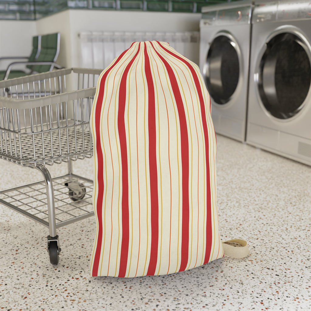 A striped laundry bag or sock holder with vertical red and cream stripes, standing on a speckled laundry room floor next to a metal shopping cart with washing machines in the background.