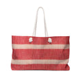 A woven tote bag with horizontal red and beige stripes, featuring two white rope handles and decorative white pom-pom accents at the top.
