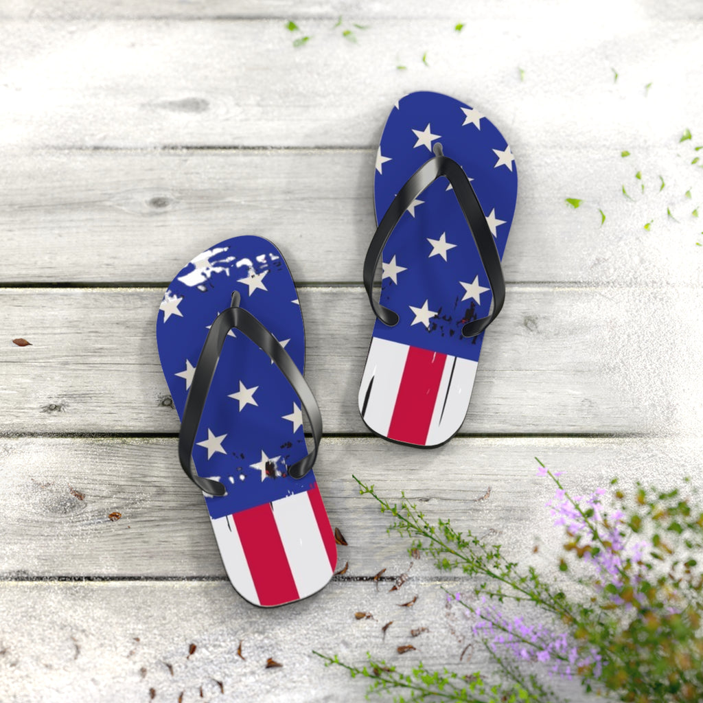 A pair of flip-flop sandals with a distressed American flag design, featuring blue straps with white stars and footbeds with red and white stripes, displayed on a light gray wooden plank surface with decorative green and purple wildflowers.