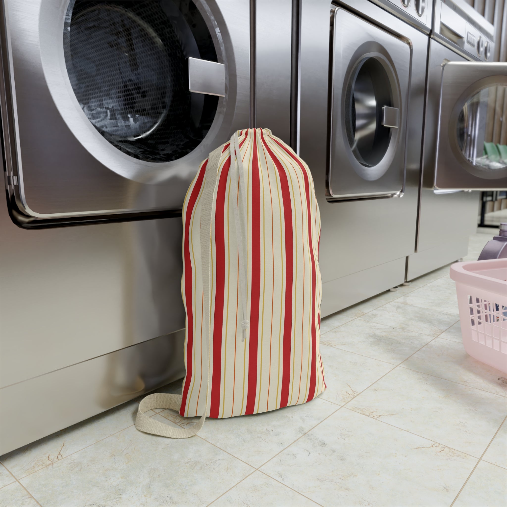 A striped fabric laundry bag with red, cream, and beige vertical stripes stands on a light tile floor in front of stainless steel washing machines.