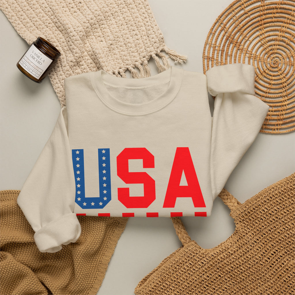 A folded cream sweatshirt with a large 'USA' graphic across the chest, styled in a flat lay with a woven blanket, a small candle jar, a wicker tray, and a tan woven bag on a light surface.