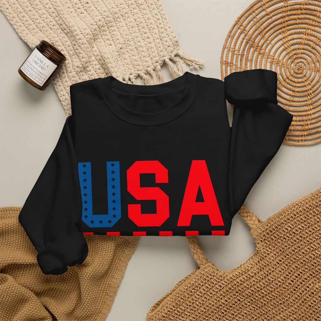 A black long-sleeve crewneck sweatshirt laid flat on a light beige surface, featuring a large 'USA' graphic across the chest in red and blue. The sweatshirt is styled with a cream knit blanket, a small amber candle jar, a woven placemat, and a tan woven bag.