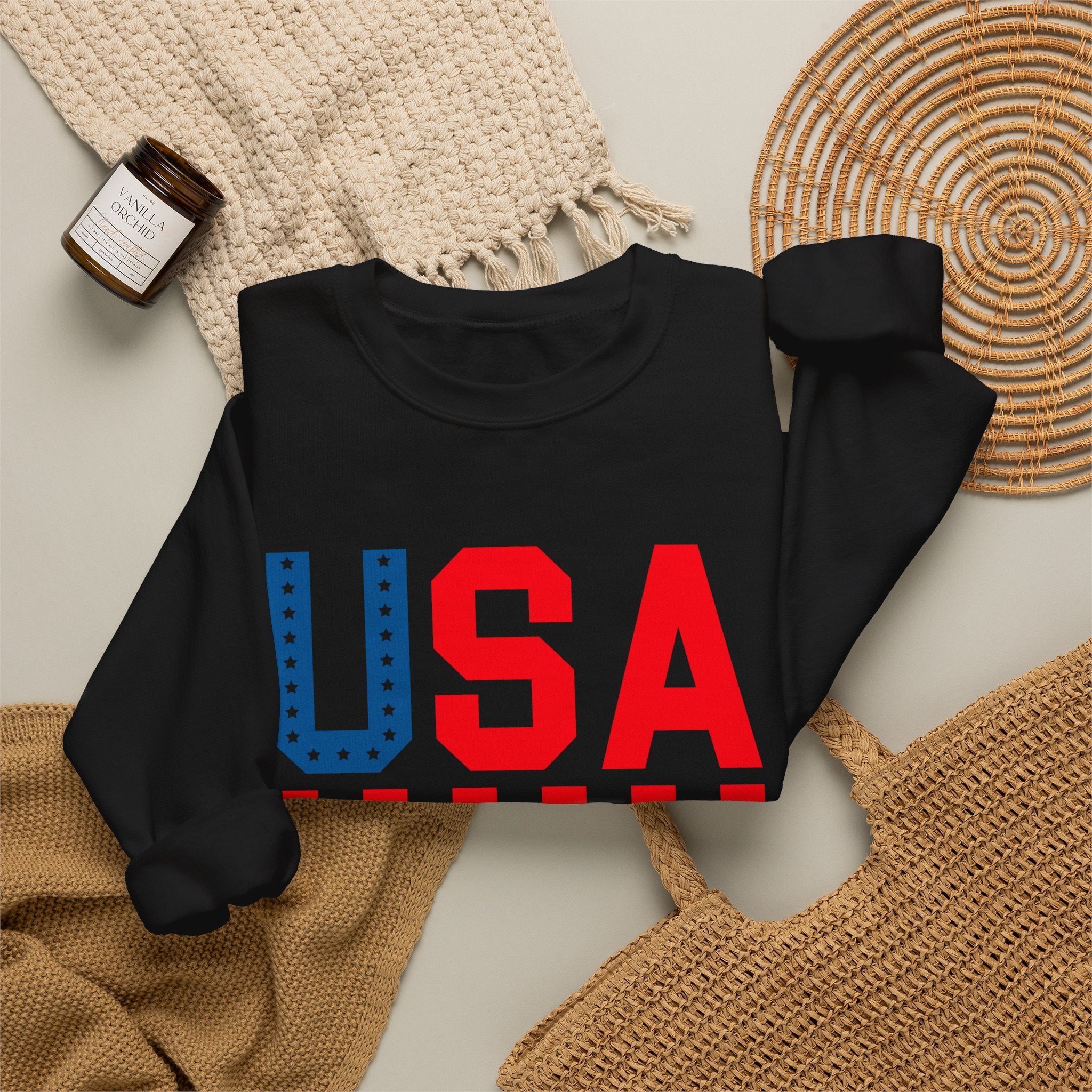 A black long-sleeve crewneck sweatshirt laid flat on a light beige surface, featuring a large 'USA' graphic across the chest in red and blue. The sweatshirt is styled with a cream knit blanket, a small amber candle jar, a woven placemat, and a tan woven bag.