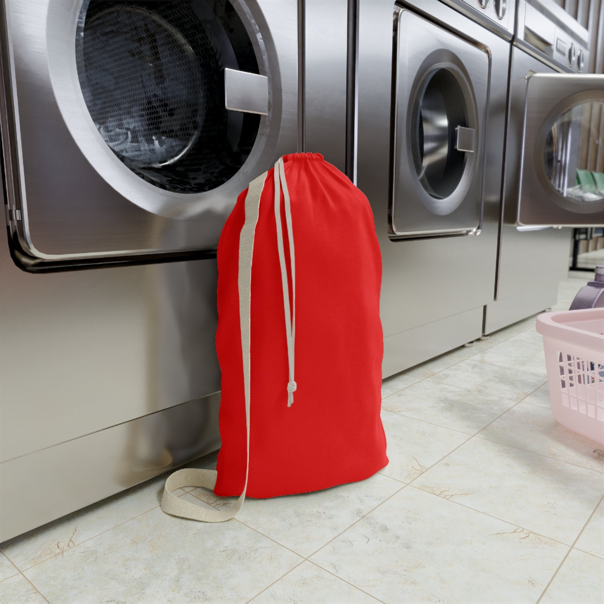 A bright red drawstring laundry bag with a beige shoulder strap and drawstrings, standing on a tiled floor in front of stainless steel washing machines.