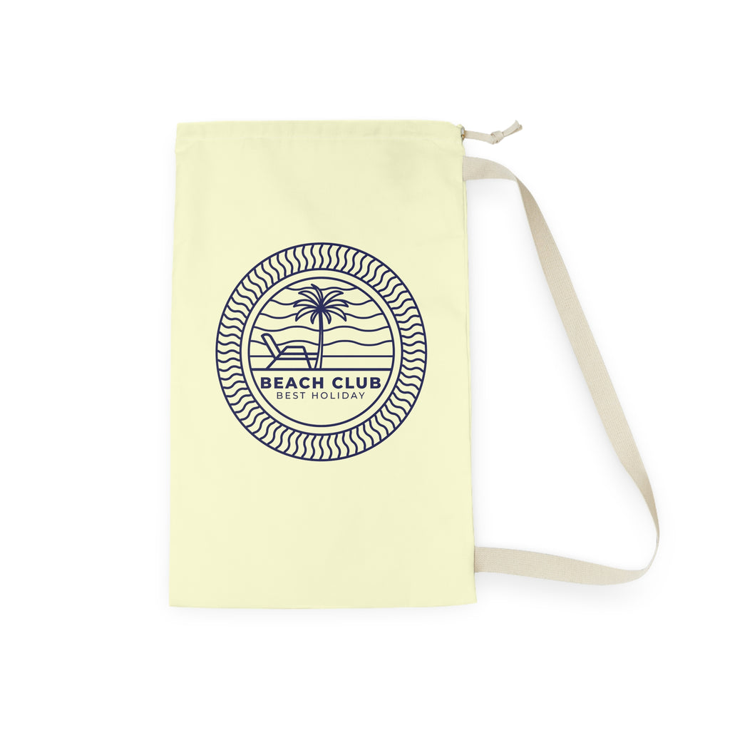 A pale yellow canvas drawstring bag with a navy blue circular graphic that reads 'BEACH CLUB EST. HOLIDAY' with a palm tree and lounge chair illustration.