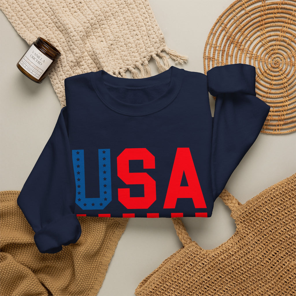 A navy blue crewneck sweatshirt with a large 'USA' graphic on the front, styled in a flat lay with a beige knit blanket, a woven placemat, a small candle jar, and a tan woven bag on a light neutral background.