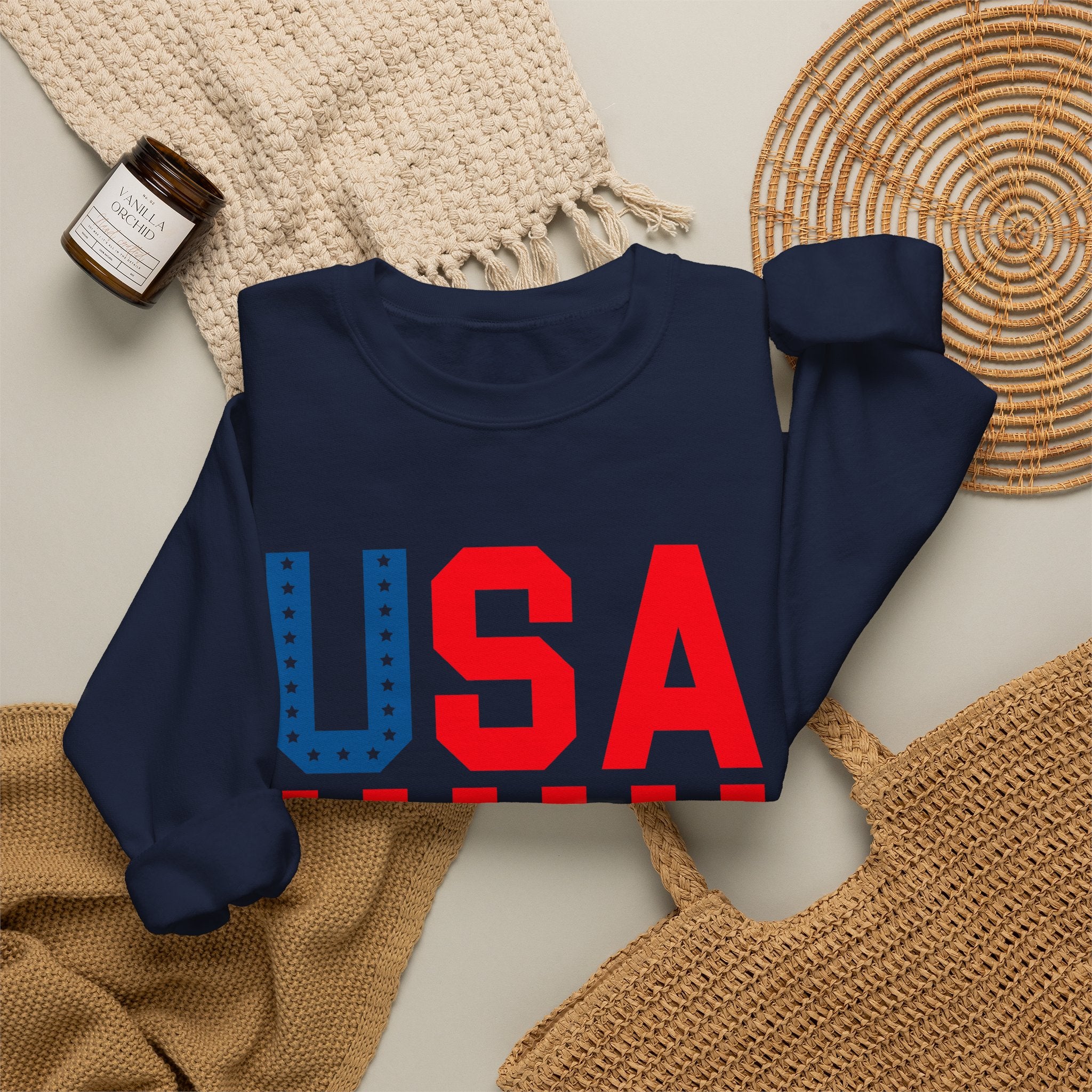 A navy blue crewneck sweatshirt with a large 'USA' graphic on the front, styled in a flat lay with a beige knit blanket, a woven placemat, a small candle jar, and a tan woven bag on a light neutral background.