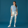 A woman modeling high-waisted leggings with a blue and yellow floral watercolor pattern on a white background, paired with a light blue t-shirt and white sneakers, set against a solid blue studio backdrop.