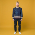 A man stands against a solid mustard yellow studio background wearing a navy blue crewneck sweatshirt with a large ship wheel graphic and horizontal red, white, and gray stripes, paired with dark jeans and white sneakers.