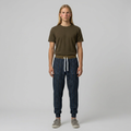 A model stands against a light gray studio background wearing an olive green short-sleeve t-shirt, dark blue patterned jogger pants with a white drawstring, and gray sneakers.