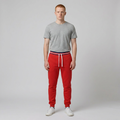 A man stands against a light gray studio background wearing bright red jogger sweatpants with a navy, white, and red striped elastic waistband and white drawstrings, paired with a heather gray t-shirt and white sneakers.