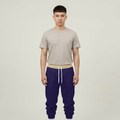 A man modeling a light beige short-sleeve t-shirt and dark purple sweatpants with a beige waistband and white drawstrings, set against a plain light gray studio background.