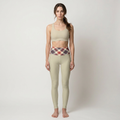 A woman stands barefoot against a light gray studio background wearing a beige sports bra and matching high-waisted leggings with a red, navy, and beige plaid waistband.