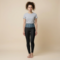 A woman stands barefoot against a plain beige studio background, wearing a light gray short-sleeve crew-neck t-shirt and black leggings with a blue geometric patterned waistband.