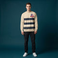 A man wearing a cream and navy blue striped crewneck sweatshirt with a red and blue crest logo on the chest, paired with dark jeans and white sneakers, standing against a dark teal studio background.
