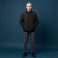 A model with platinum blonde hair stands against a dark blue studio background wearing a black quilted puffer jacket with small gold dot details, dark gray jeans, and black shoes.