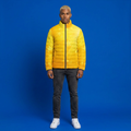 A man wearing a bright yellow quilted puffer jacket with a black zipper, paired with dark gray jeans and white sneakers, standing against a solid blue studio background.