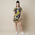 A woman modeling a short-sleeve shift dress with an abstract organic pattern in olive green, black, and white, paired with white sneakers, against a solid beige studio background.