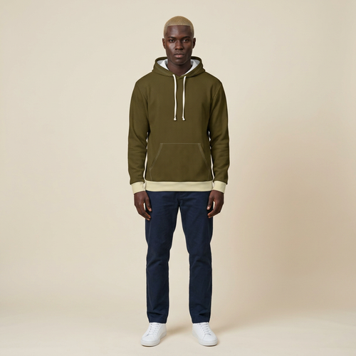 A man stands facing forward wearing an olive green pullover hoodie with cream drawstrings and ribbed trim, paired with navy pants and white sneakers, against a plain beige studio background.