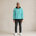 A model stands facing forward wearing a turquoise hoodie with a black hood and black ribbed cuffs and hem, paired with dark gray jeans and white sneakers, against a textured beige studio backdrop.