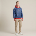 A man modeling a blue and white striped hoodie with red trim, paired with beige pants and white sneakers, against a solid beige studio background.