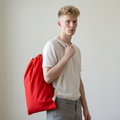 A young man with blonde hair wearing a white t-shirt and gray trousers, carrying a bright red drawstring backpack over his shoulder against a plain light gray background.