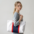 A woman wearing a gray shirt carries a large white canvas tote bag with a navy blue center panel and red side stripes, set against a plain light gray wall.
