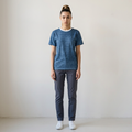 A woman stands facing forward in a studio setting, wearing a textured blue short-sleeve crewneck t-shirt, dark gray slim-fit pants, and white sneakers against a plain off-white background.