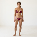 A woman modeling a two-piece string bikini in deep burgundy with light pink trim, standing against a plain white studio background.