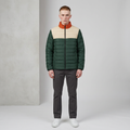 A man wearing a color-blocked puffer jacket with dark green, beige, and rust-orange panels, paired with dark gray trousers and white sneakers, standing against a light gray studio background.