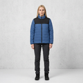 A woman stands facing forward wearing a color-blocked puffer jacket with a blue body and black yoke, paired with dark pants and black boots, against a light gray textured studio wall.