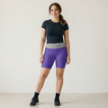 A woman modeling purple athletic biker shorts with a gray patterned waistband, paired with a dark navy t-shirt, black socks, and black sneakers, standing against a plain off-white studio background.