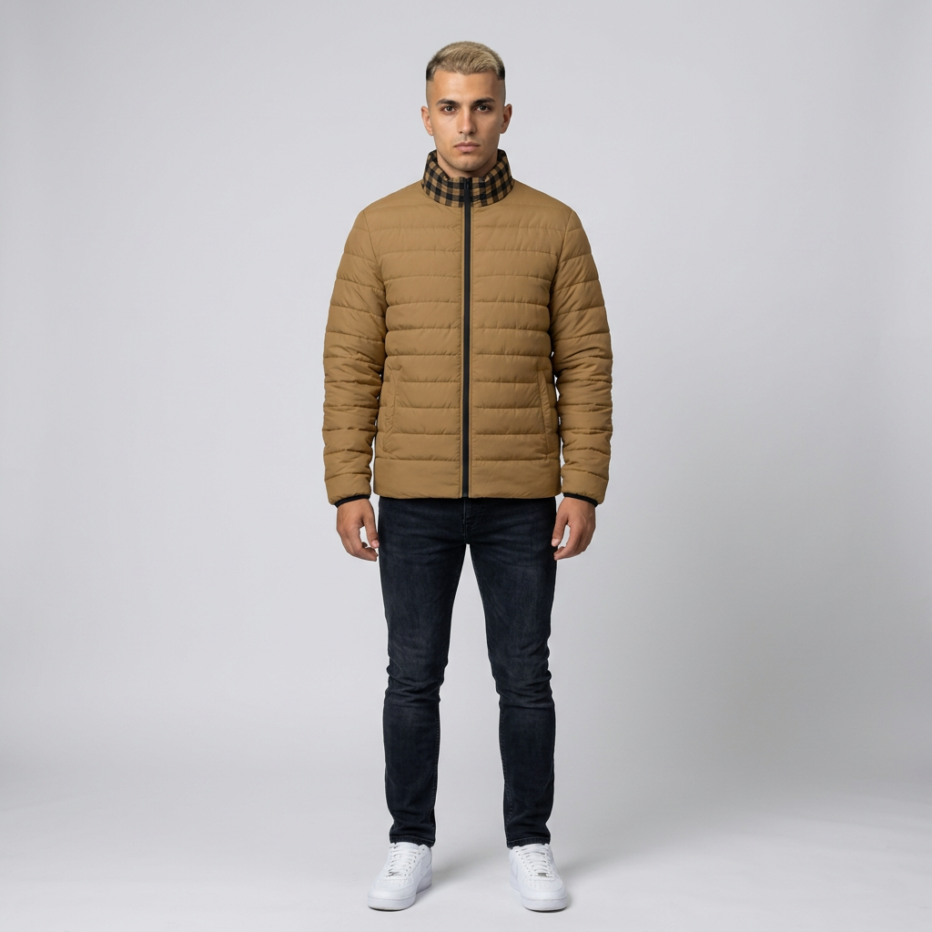 A man wearing a tan quilted puffer jacket with a black and white checkered high collar, paired with dark blue jeans and white sneakers, standing against a light gray studio background.