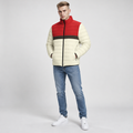 A man modeling a color-blocked puffer jacket with cream, red, and dark brown horizontal color blocks, paired with blue jeans and white sneakers against a light gray studio background.