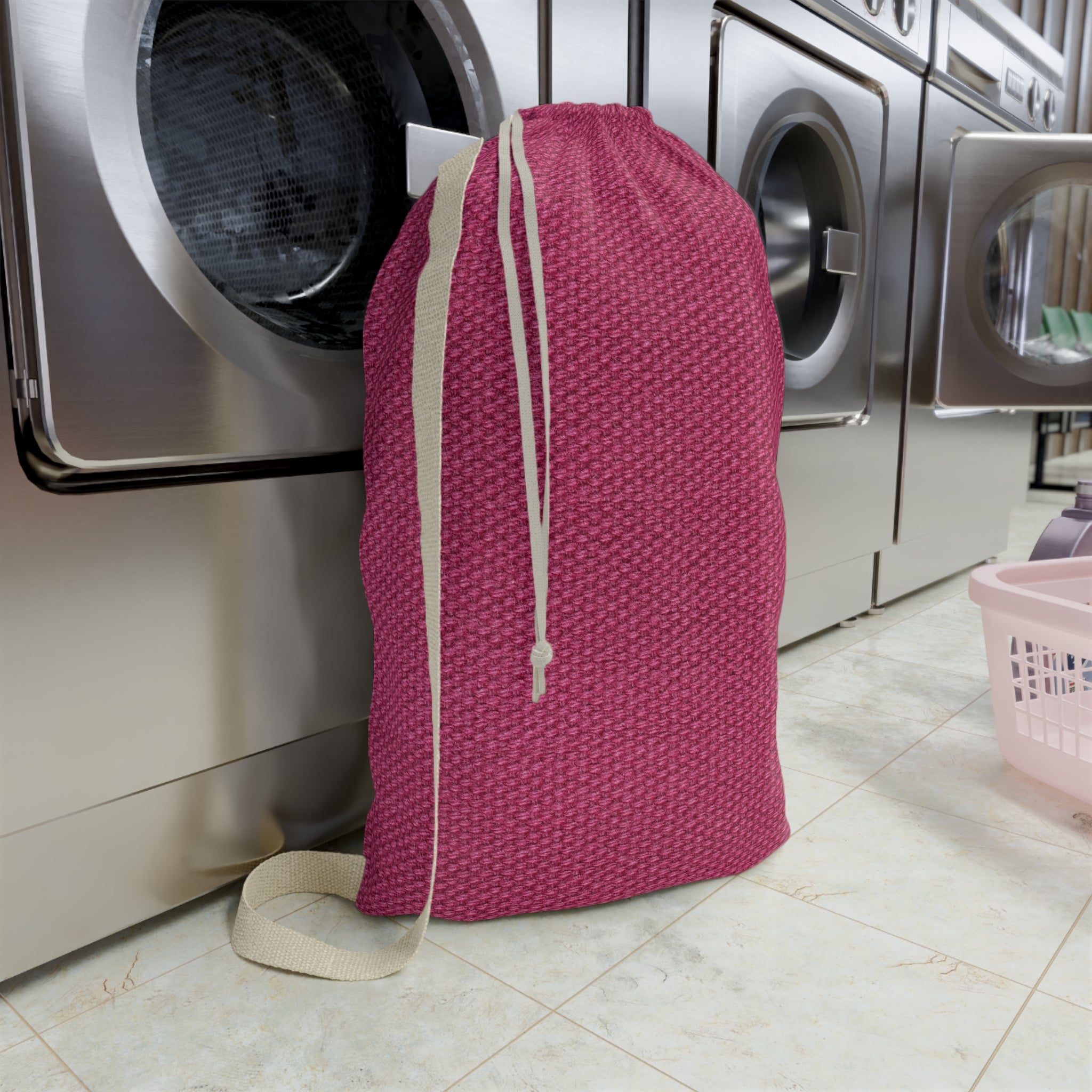 A pink textured drawstring laundry bag with beige straps stands in front of stainless steel washing machines on a tiled floor.