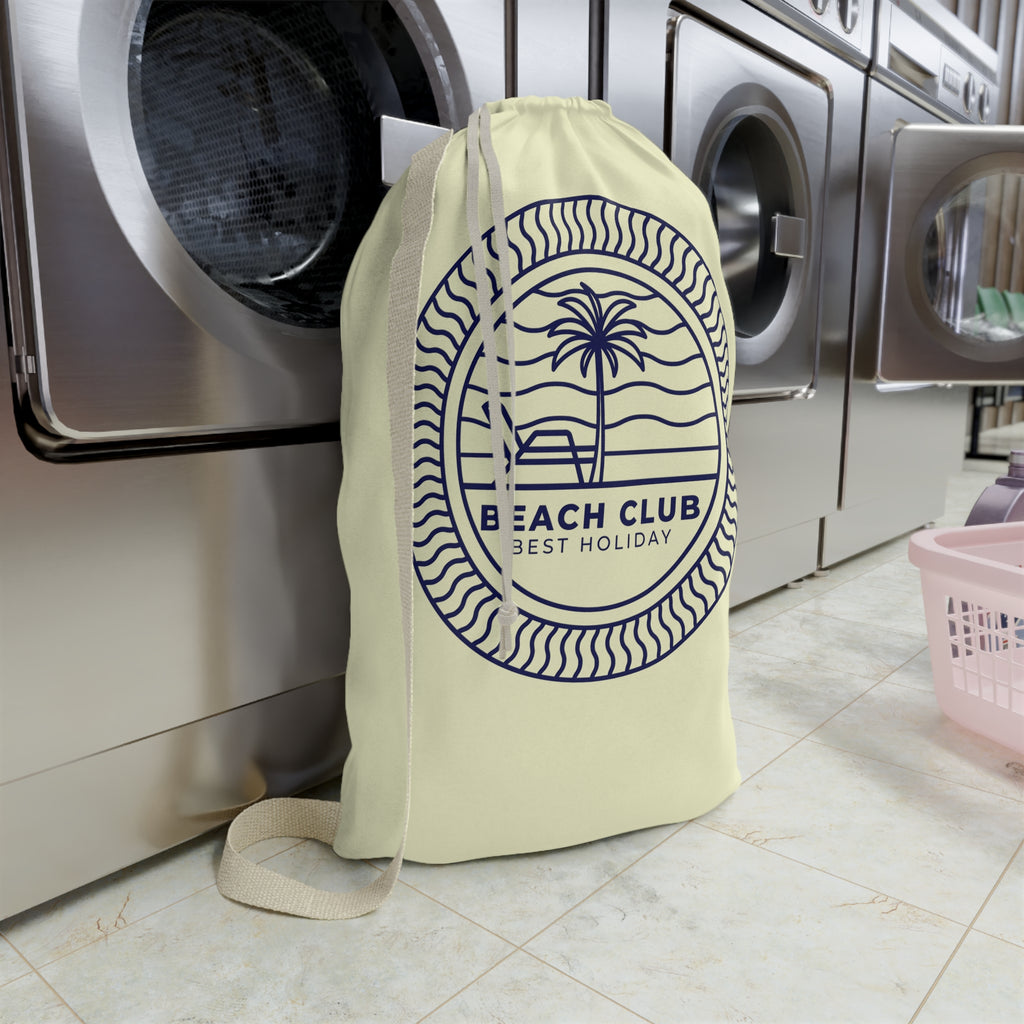 A cream-colored drawstring laundry bag with a navy blue 'Beach Club Best Holiday' palm tree graphic, standing in front of stainless steel washing machines on a tiled floor.