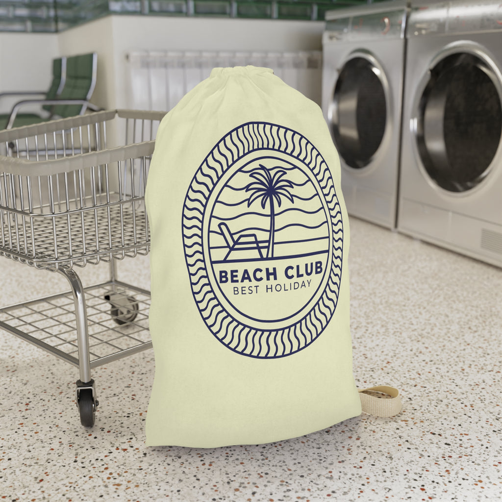 A cream-colored drawstring laundry bag with a navy blue 'BEACH CLUB BEST HOLIDAY' palm tree graphic, standing on a speckled floor next to a metal cart with washing machines in the background.