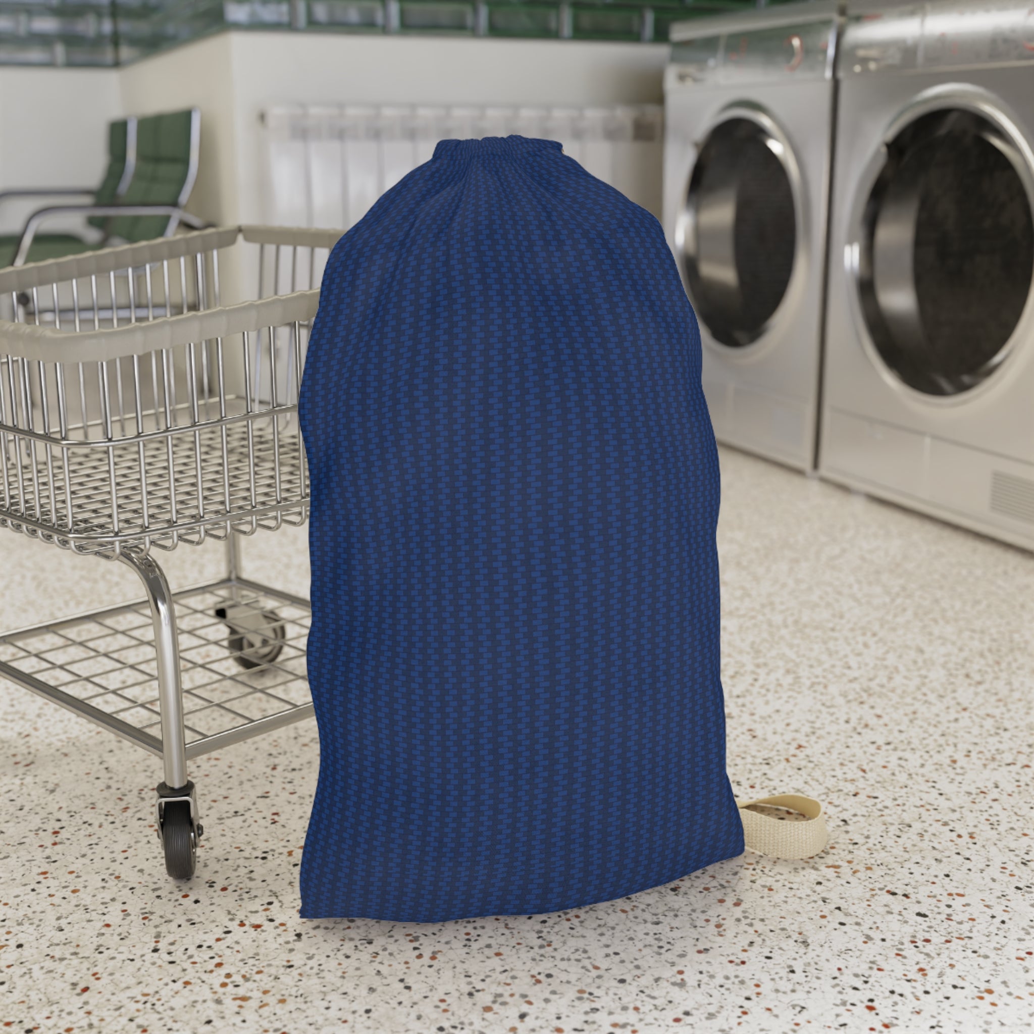 A large blue drawstring laundry bag with a subtle geometric pattern, standing on a speckled floor next to a metal laundry cart in a laundromat with washing machines in the background.